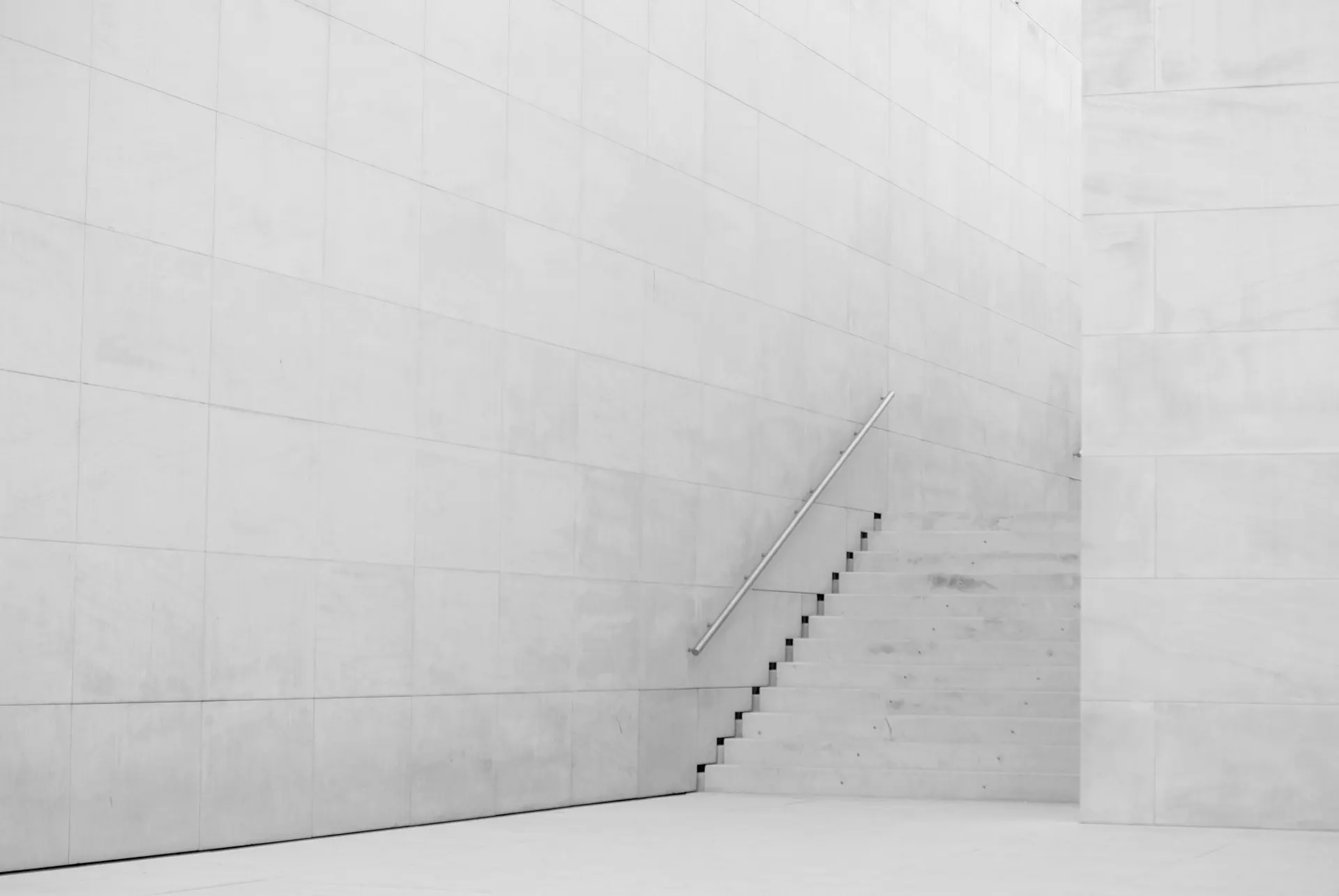 Una escalera de hormigón blanco que sube junto a un muro desnudo, con luz cenital